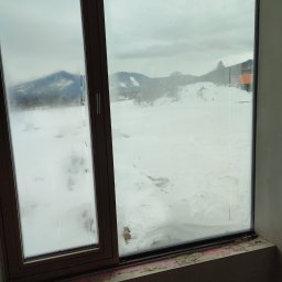 Tynki Spider - Widok z okna na zaśnieżony krajobraz górski. Okno z brązową ramą, częściowo zaparowane, z widocznymi śladami prac wykończeniowych na parapecie.