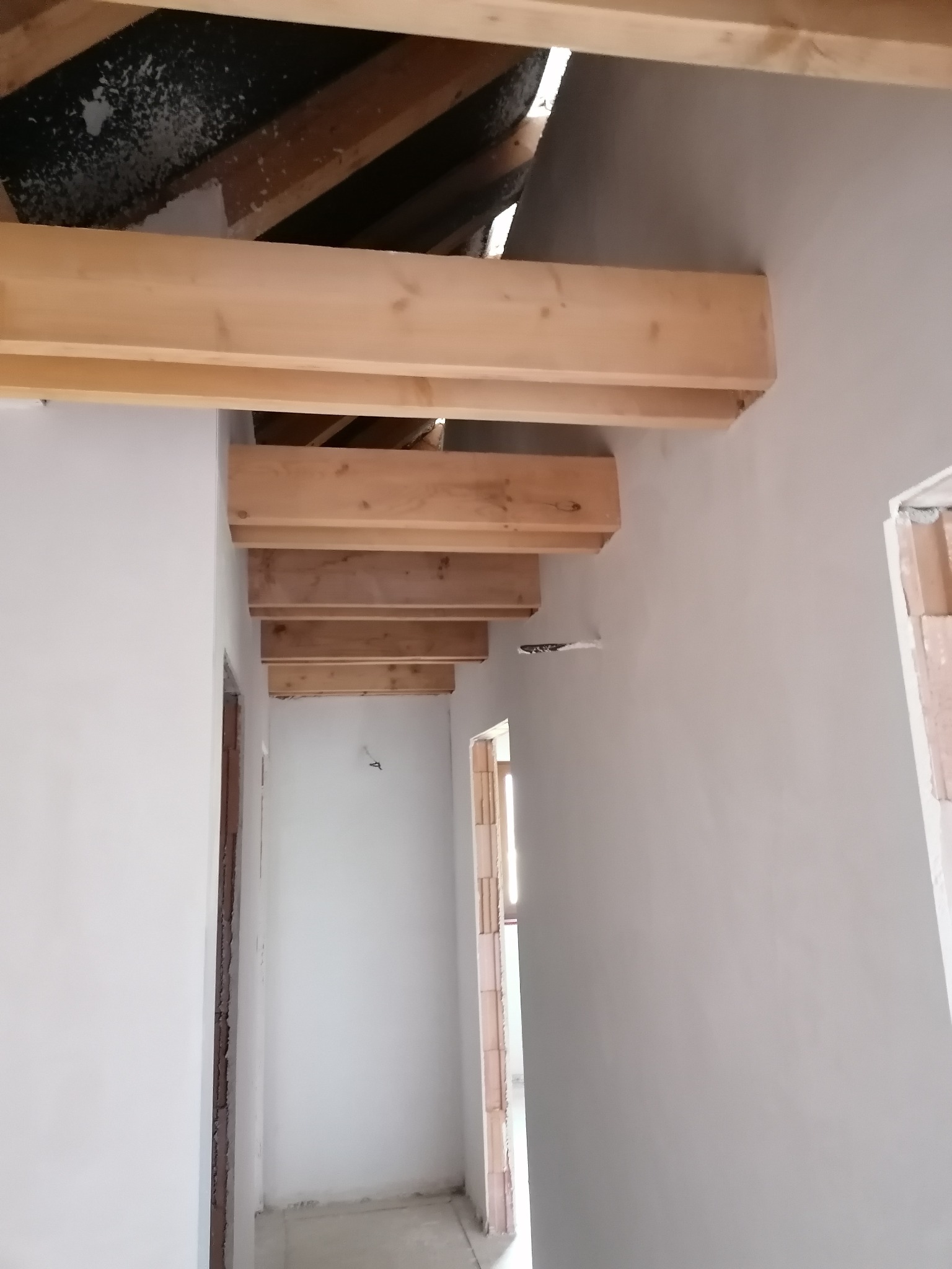 Poddasze po gładzi, z widocznymi drewnianymi belkami stropowymi. Ściany gładkie, pomalowane na jasny kolor. Widoczne fragmenty okien i drzwi w stanie surowym.