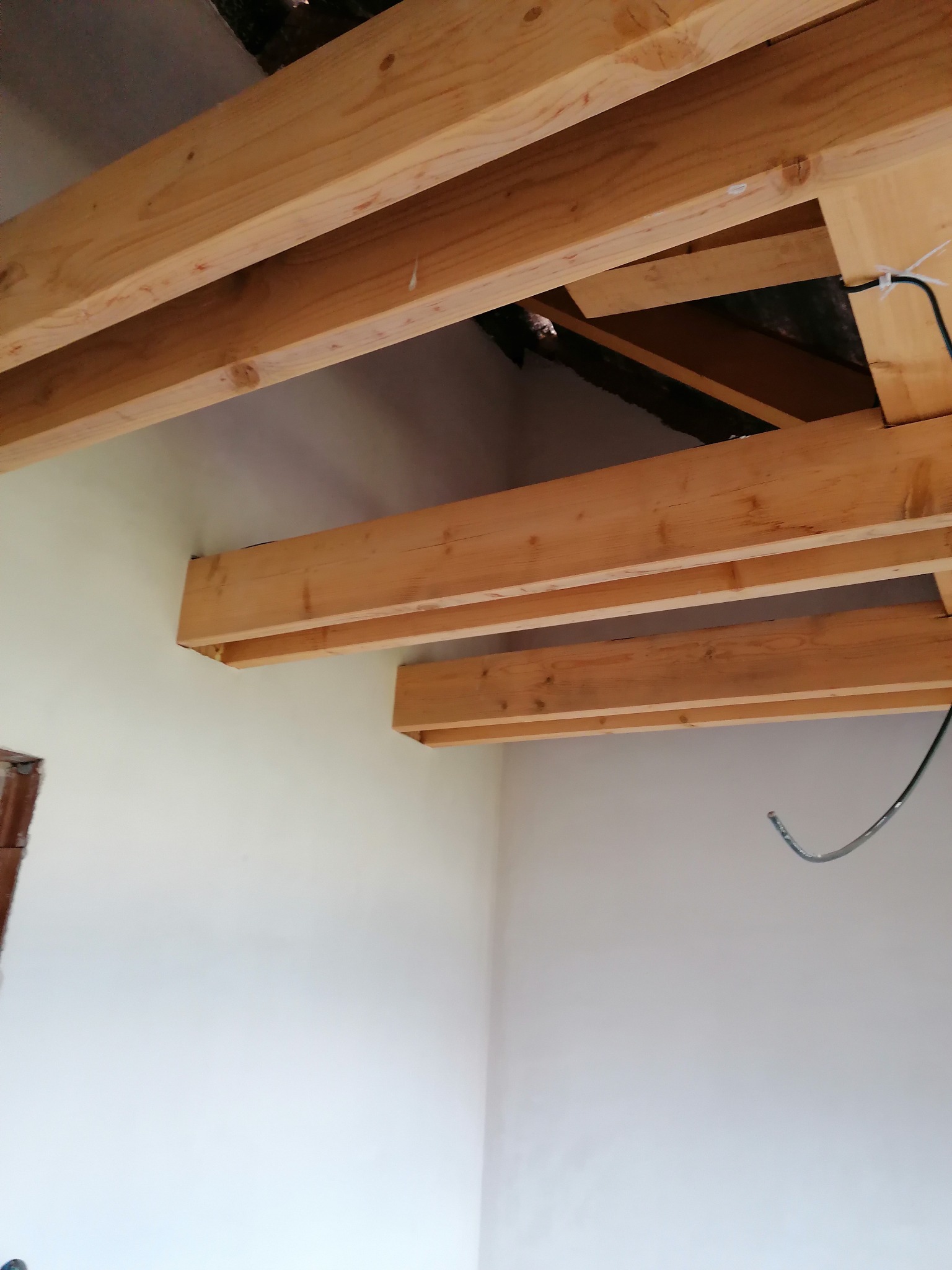 Drewniana konstrukcja poddasza z widocznymi belkami i fragmentem instalacji elektrycznej na tle białej ściany. Ujęcie z dołu, ukazujące surowy stan przed wykończeniem.