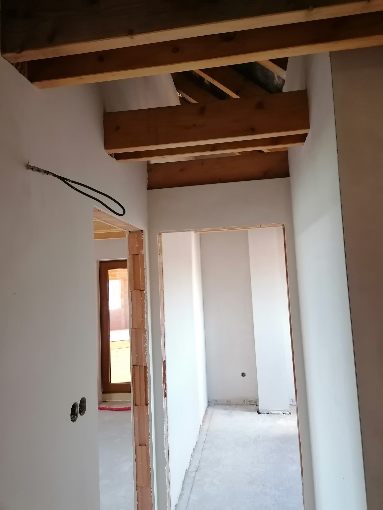 Wnętrze w trakcie wykończenia: białe ściany po gładzi, widoczne drewniane belki stropowe, instalacja elektryczna i surowa ościeżnica drzwiowa z cegły.