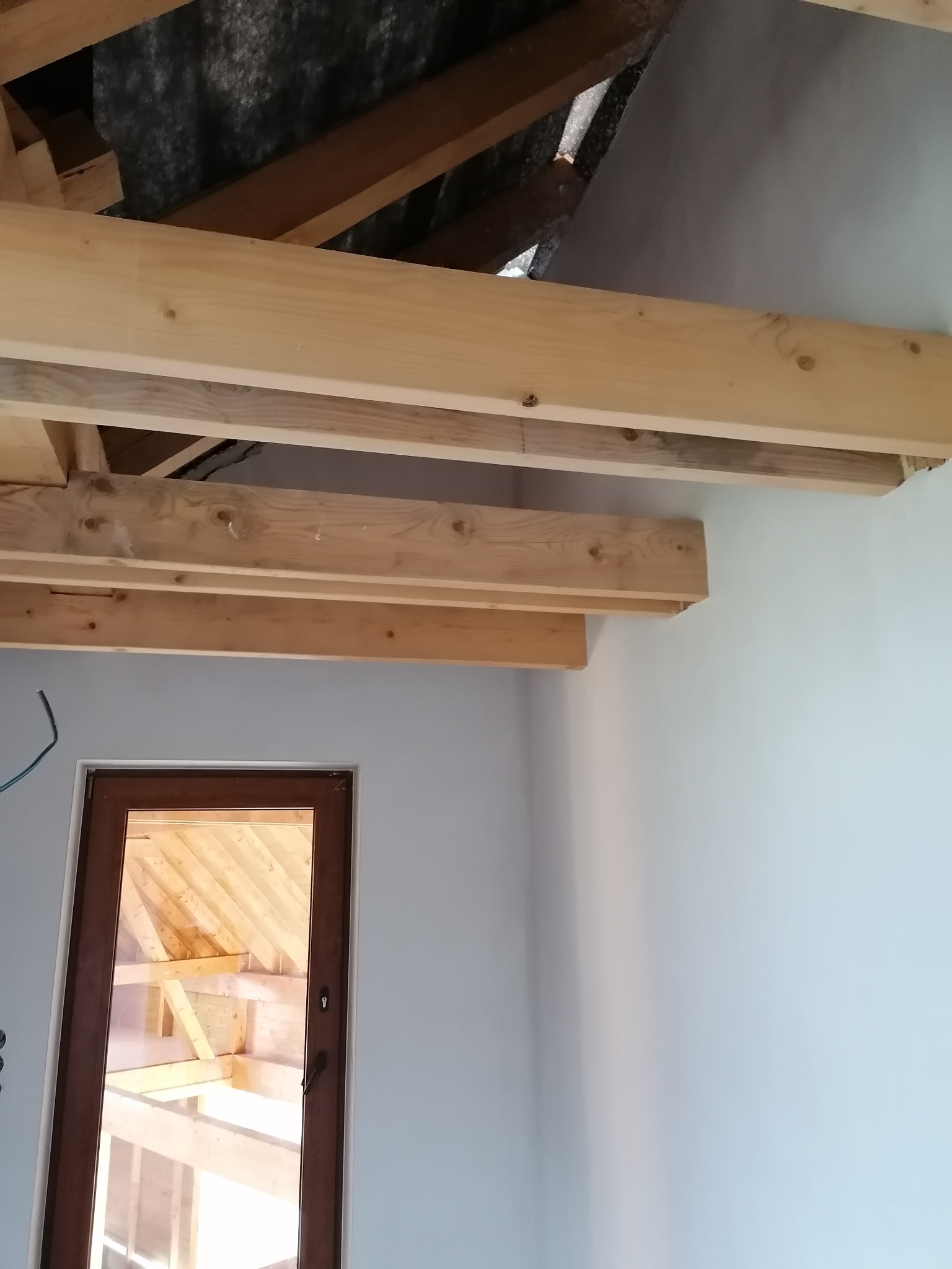 Poddasze w trakcie ocieplania: drewniana konstrukcja stropu, widoczna izolacja, fragment ściany i okno. Prace wykończeniowe w jasnym wnętrzu.