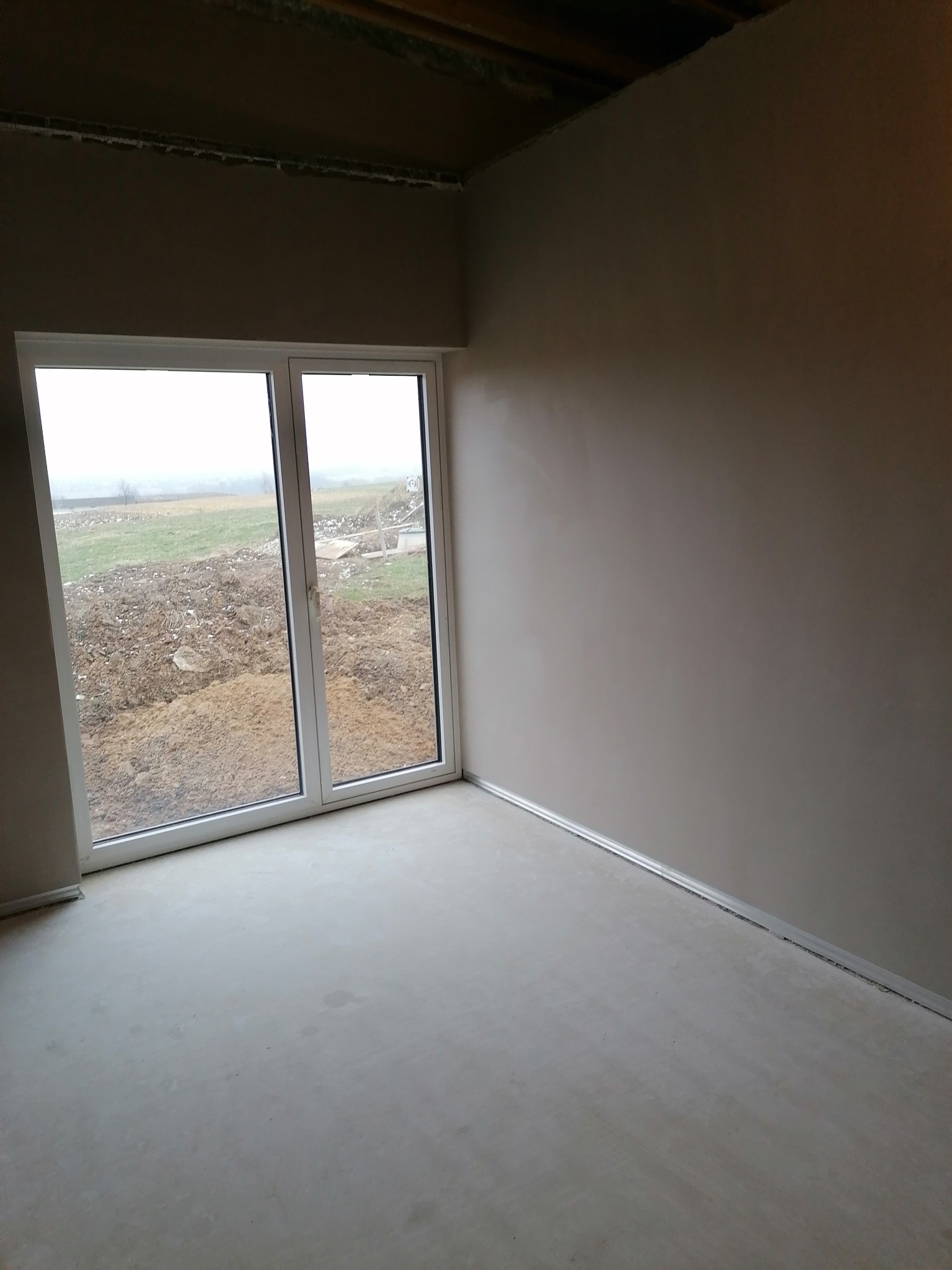 Wnętrze pokoju po gładziowaniu ścian i wylewce podłogi, z widokiem na zewnątrz przez białe okno balkonowe. Ściany w odcieniach szarości, surowy stan wykończenia.
