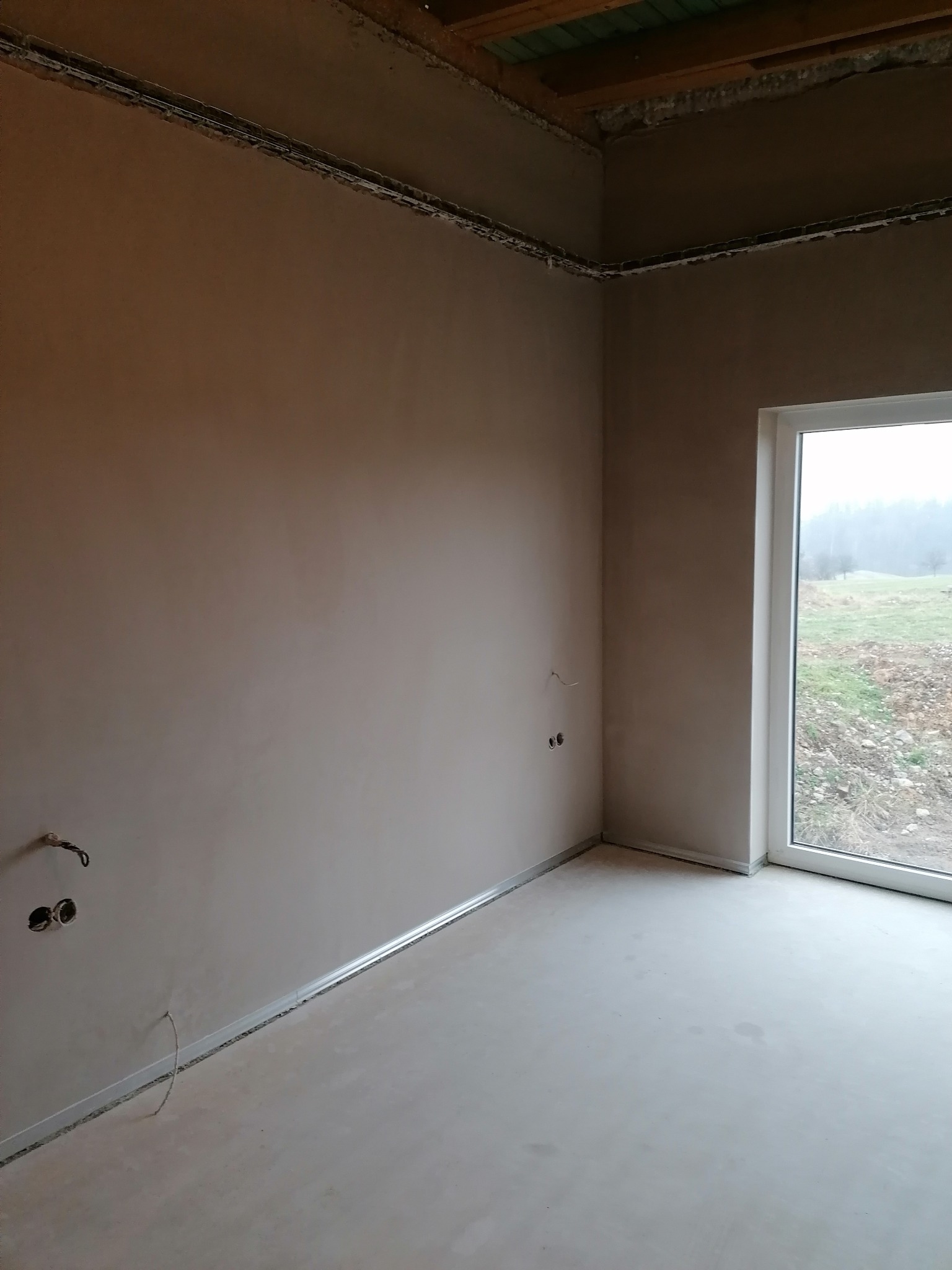 Wnętrze pokoju po tynkowaniu ścian i wylewce podłogi, widoczne puszki elektryczne i listwy przypodłogowe. Przez okno widok na zewnątrz.