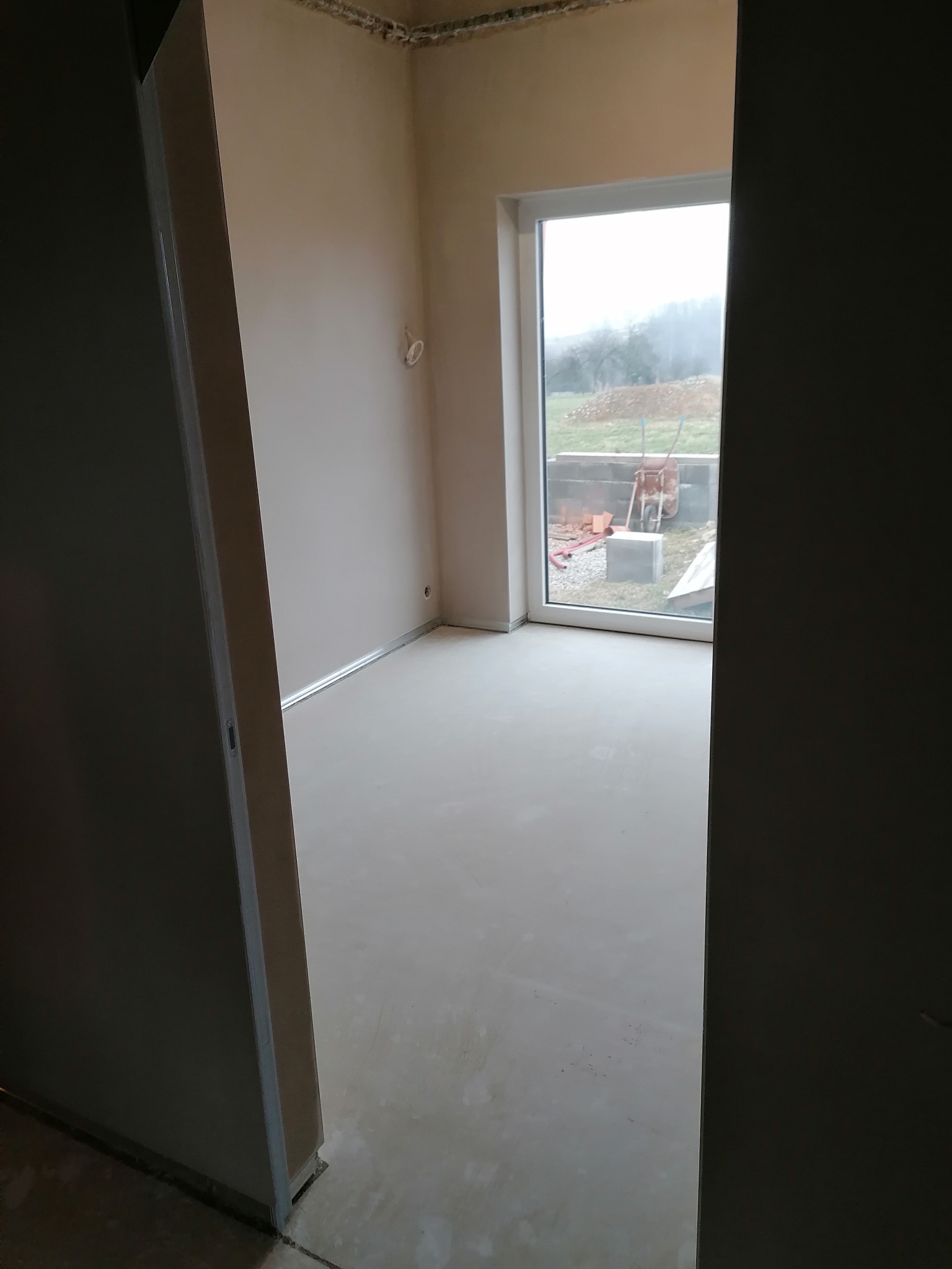Wnętrze pokoju po wylaniu posadzki, ściany przygotowane do malowania. Widoczne okno z widokiem na zewnątrz i fragment drzwi.