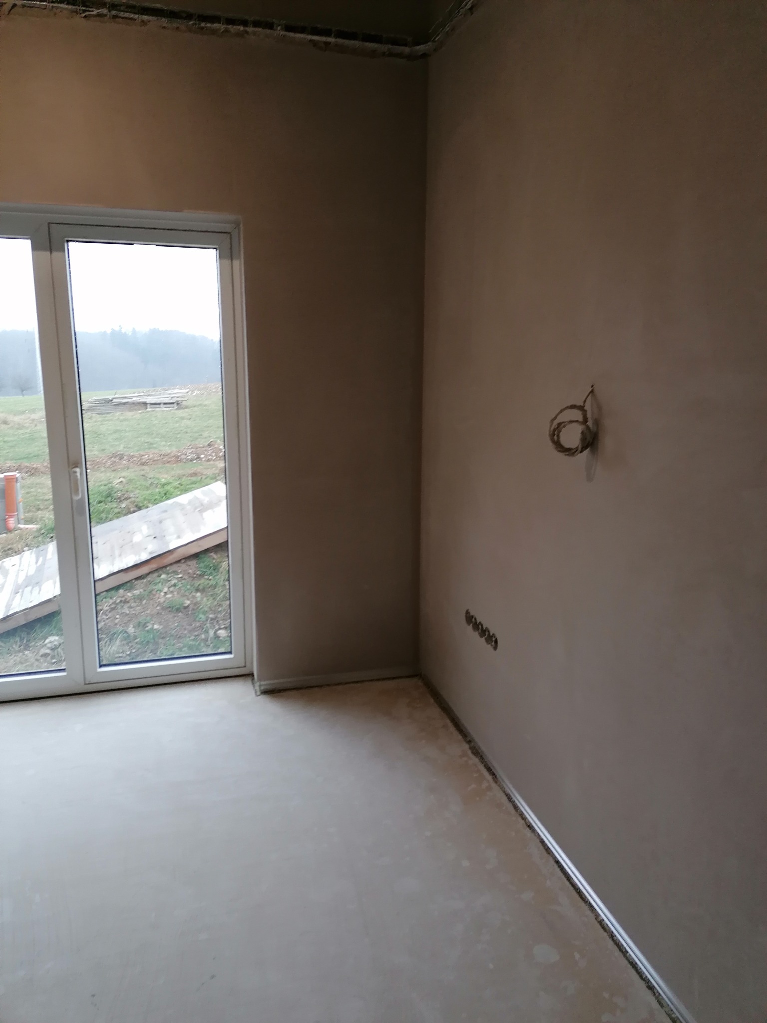 Wnętrze pokoju po nałożeniu gładzi, widoczne gniazdka elektryczne i wyprowadzone przewody. Ściany w kolorze beżowym, okno balkonowe z widokiem na zewnątrz.