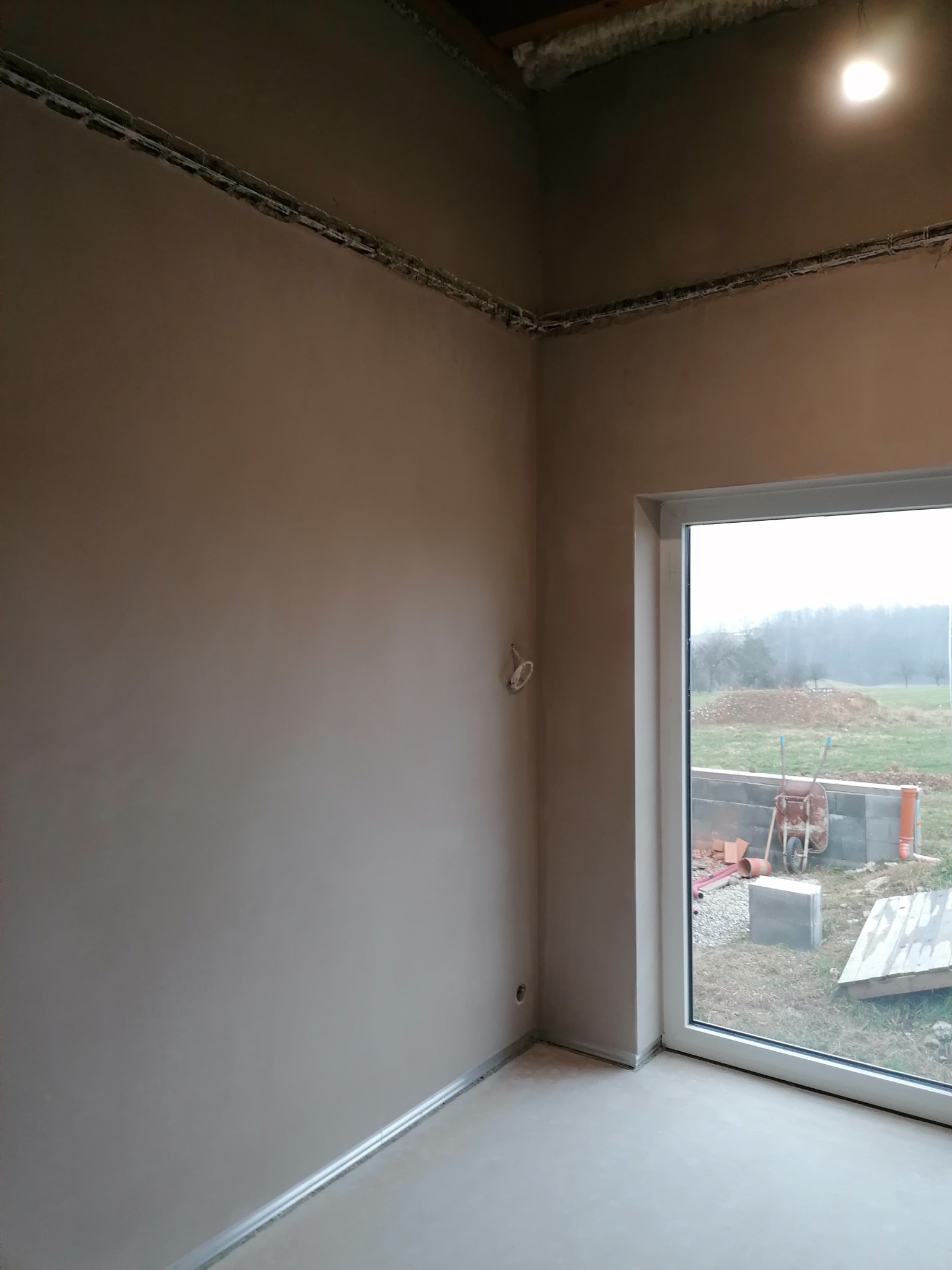 Wnętrze po tynkowaniu: gładka, beżowa ściana z widocznymi przewodami elektrycznymi pod sufitem, okno z widokiem na zewnątrz i fragment podłogi. Minimalistyczny efekt.