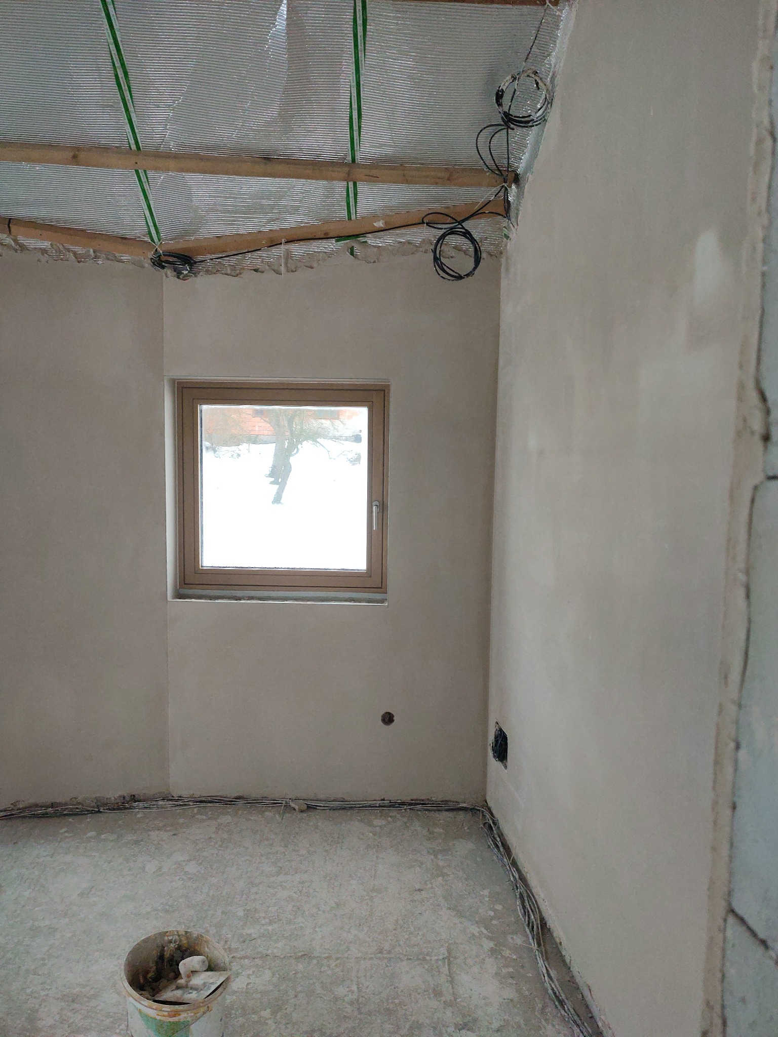 Pokój po tynkowaniu: świeże, jasne ściany, okno z widokiem na zimowy krajobraz, widoczne przewody elektryczne i wiadro z narzędziami na podłodze.