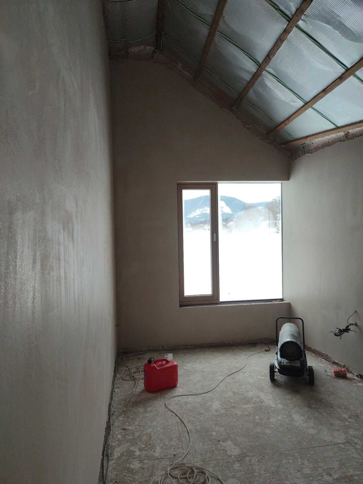 Pokój po tynkowaniu ścian, widoczne świeże tynki, okno z widokiem na zaśnieżone góry, nagrzewnica i kanister. Poddasze z izolacją.