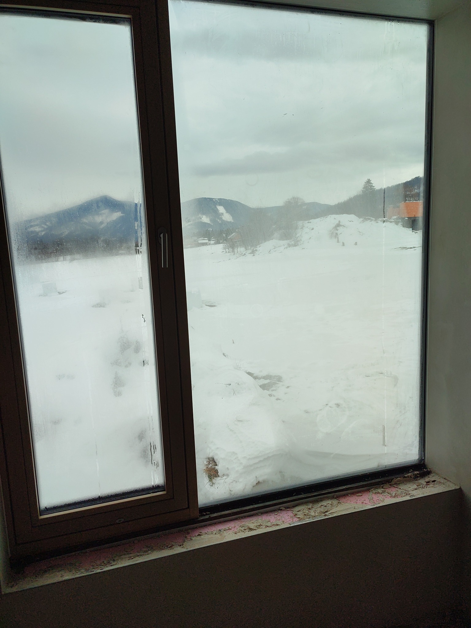 Widok z okna na zaśnieżony krajobraz górski. Okno z brązową ramą, częściowo zaparowane, z widocznymi śladami prac wykończeniowych na parapecie.