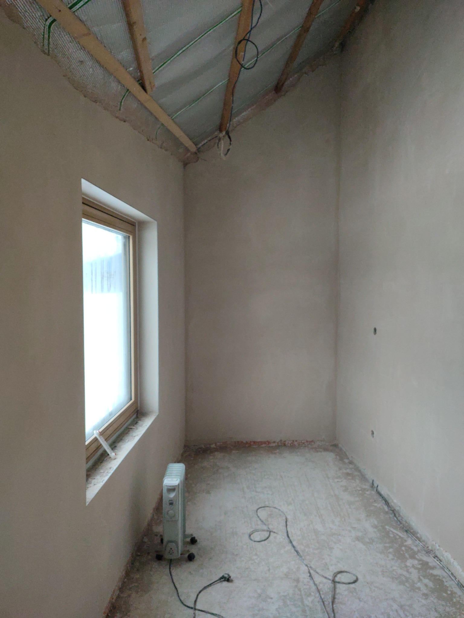 Pokój po tynkowaniu ścian, widoczne gładkie, szare ściany, okno z grzejnikiem i fragment poddasza z izolacją. Kabel elektryczny na podłodze.