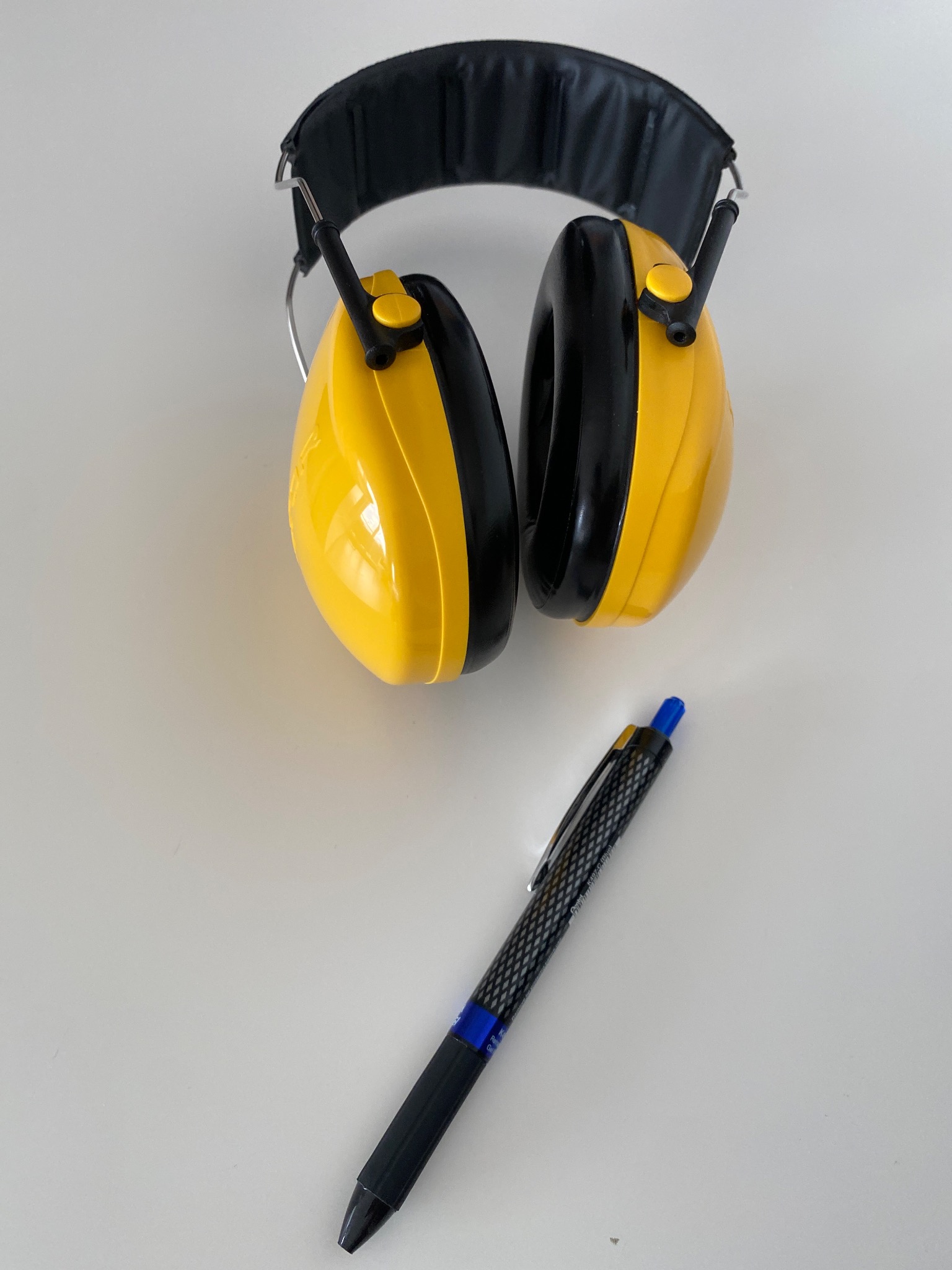 Żółto-czarne słuchawki ochronne i czarny długopis na białym tle. Słuchawki leżą na górze, długopis poniżej, sugerując przygotowanie do pracy na budowie.