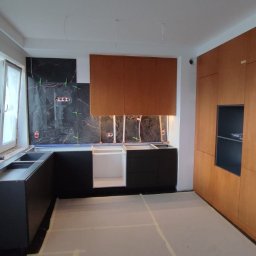 Serhii Kalataliuk - Częściowo zmontowana kuchnia z czarnymi szafkami dolnymi i drewnianymi górnymi. Widoczne płytki imitujące marmur i przygotowane instalacje elektryczne.
