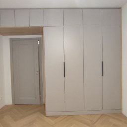 Serhii Kalataliuk - Jasnoszara szafa na wymiar z górnymi szafkami, czarne uchwyty, parkiet w jodełkę. Widoczne niedokończone gniazdka elektryczne. Minimalistyczny design.