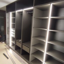 Serhii Kalataliuk - Nowoczesna szafa wnękowa z szarymi frontami i oświetleniem LED, widoczne półki i szuflady. Elementy montażowe na podłodze sugerują etap końcowy instalacji.