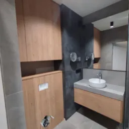 Nowoczesna łazienka z drewnianą zabudową meblową, szarą płytką i okrągłą umywalką. Widoczne lustra i oświetlenie punktowe.