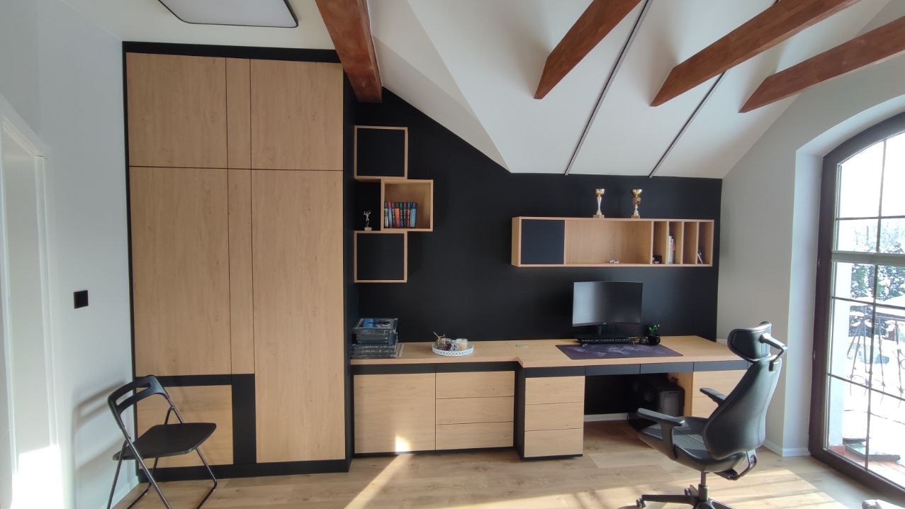 Pokój na poddaszu z zabudową meblową: szafa, biurko z szufladami i półki w odcieniach drewna z czarnymi akcentami. Widoczne drewniane belki stropowe i okno.