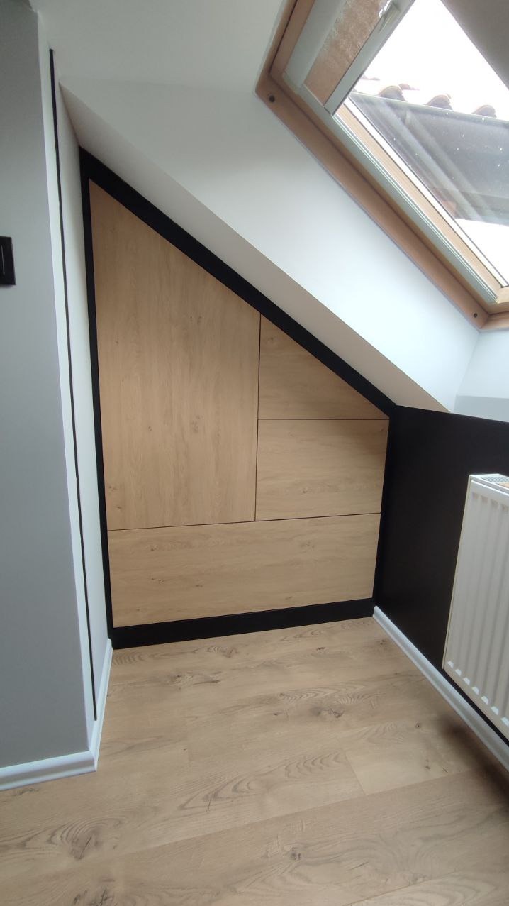 Drewniana szafa wnękowa na poddaszu, z czarnymi obramowaniami, dopasowana do skosu dachu. Widoczne panele podłogowe i okno dachowe.