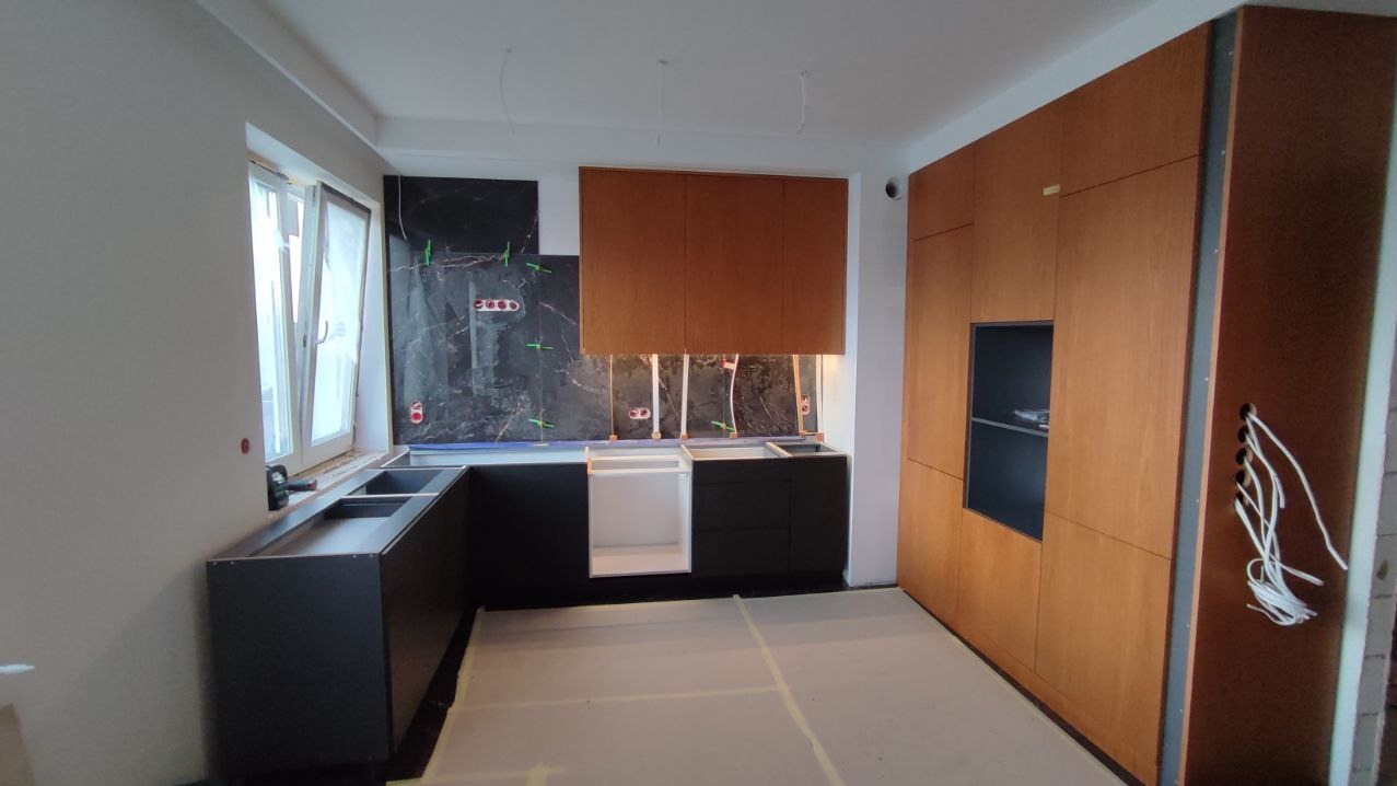 Częściowo zmontowana kuchnia z czarnymi szafkami dolnymi i drewnianymi górnymi. Widoczne płytki imitujące marmur i przygotowane instalacje elektryczne.