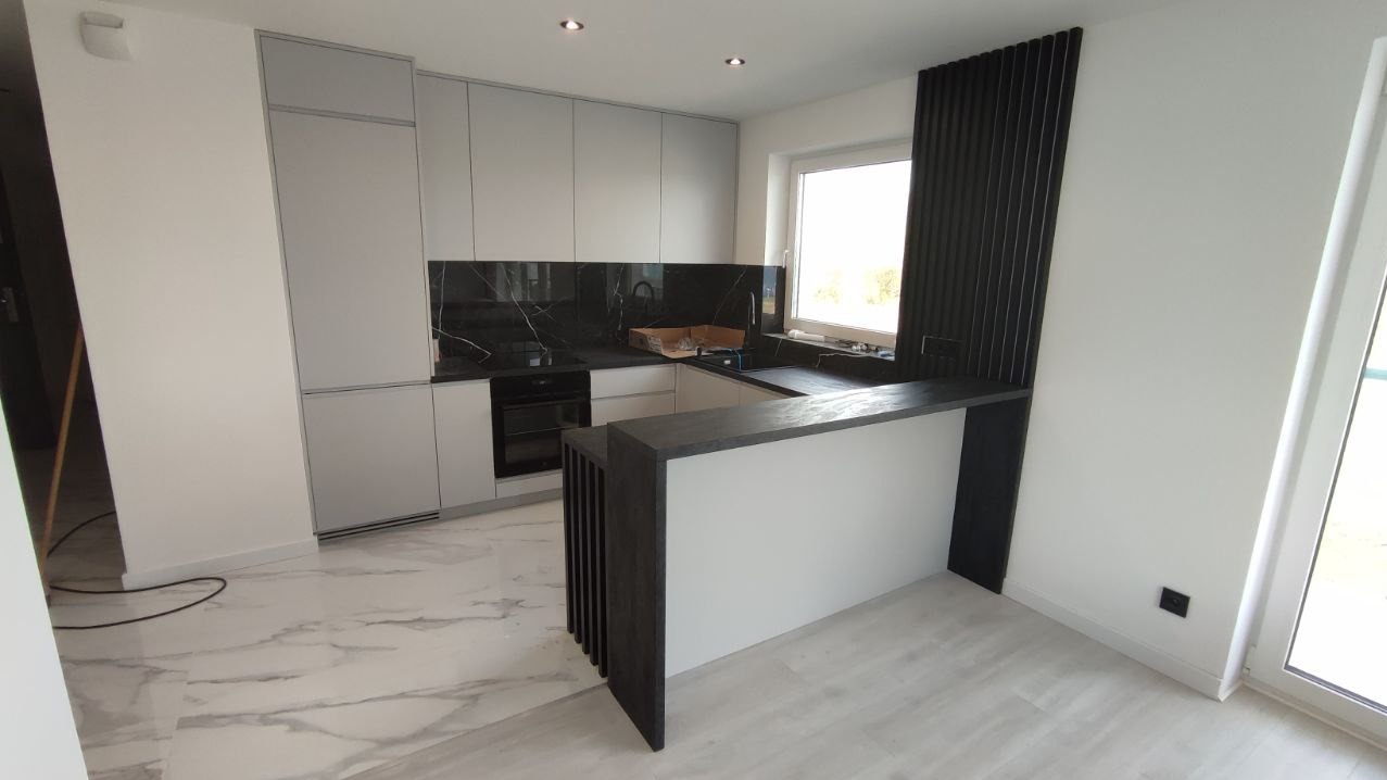 Nowoczesna kuchnia z szarymi szafkami, czarnym blatem i marmurową podłogą. Wyspa kuchenna z drewnianymi lamelami dodaje elegancji. Jasne wnętrze z oknem.