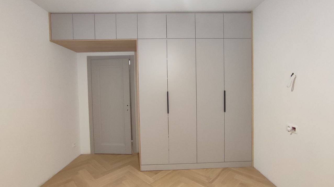 Jasnoszara szafa na wymiar z górnymi szafkami, czarne uchwyty, parkiet w jodełkę. Widoczne niedokończone gniazdka elektryczne. Minimalistyczny design.