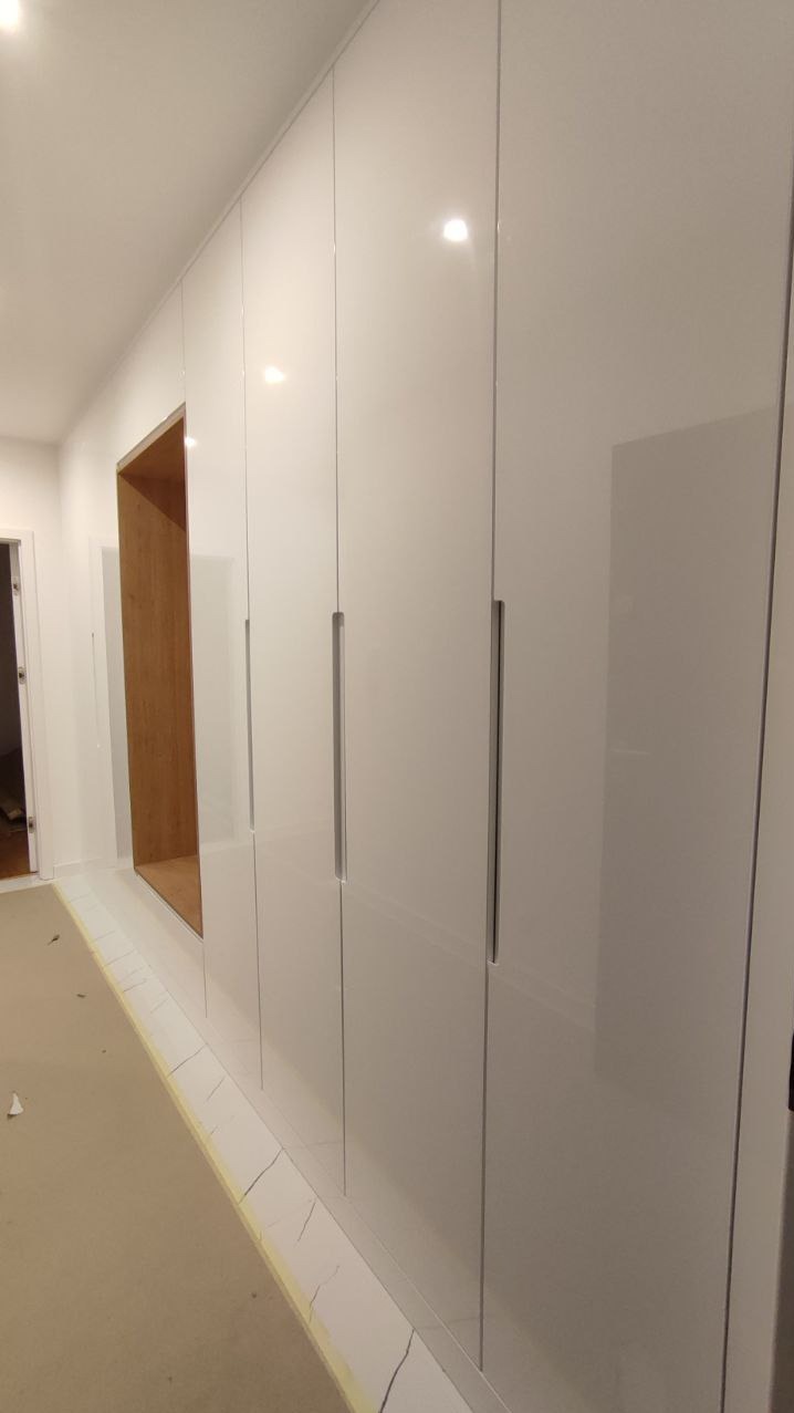 Biała szafa wnękowa z połyskiem i wnęką z drewnianym wykończeniem. Minimalistyczny design, widoczne detale wykończeniowe przy podłodze.