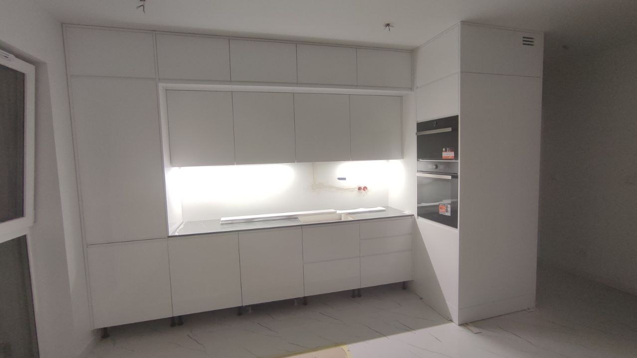 Minimalistyczna, biała kuchnia na wymiar z podświetleniem LED pod szafkami wiszącymi. Widoczne zabudowane piekarniki i marmurowa podłoga.