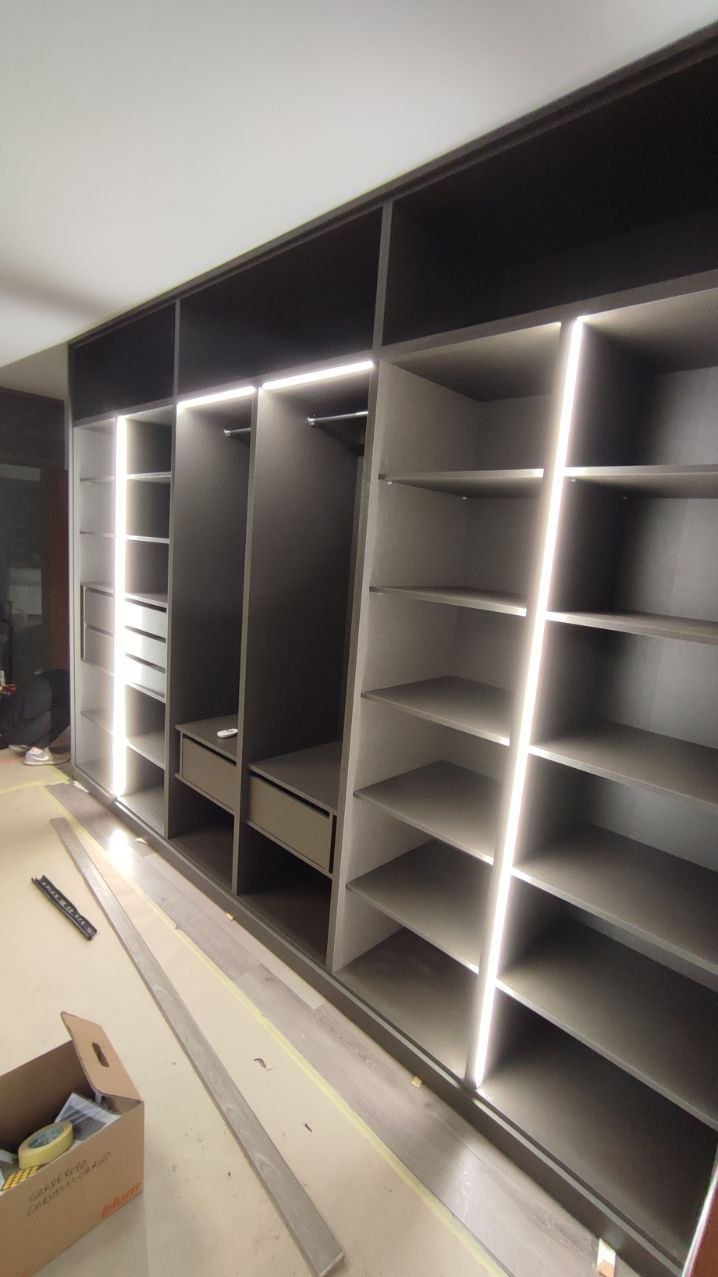 Nowoczesna szafa wnękowa z szarymi frontami i oświetleniem LED, widoczne półki i szuflady. Elementy montażowe na podłodze sugerują etap końcowy instalacji.