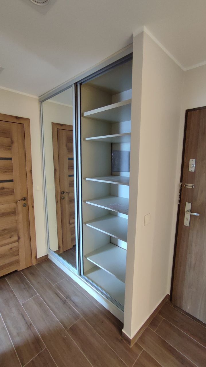 Biała szafa wnękowa z lustrzanymi drzwiami przesuwnymi, widok z boku, na tle drewnianych drzwi i podłogi. Realizacja w Katowicach.