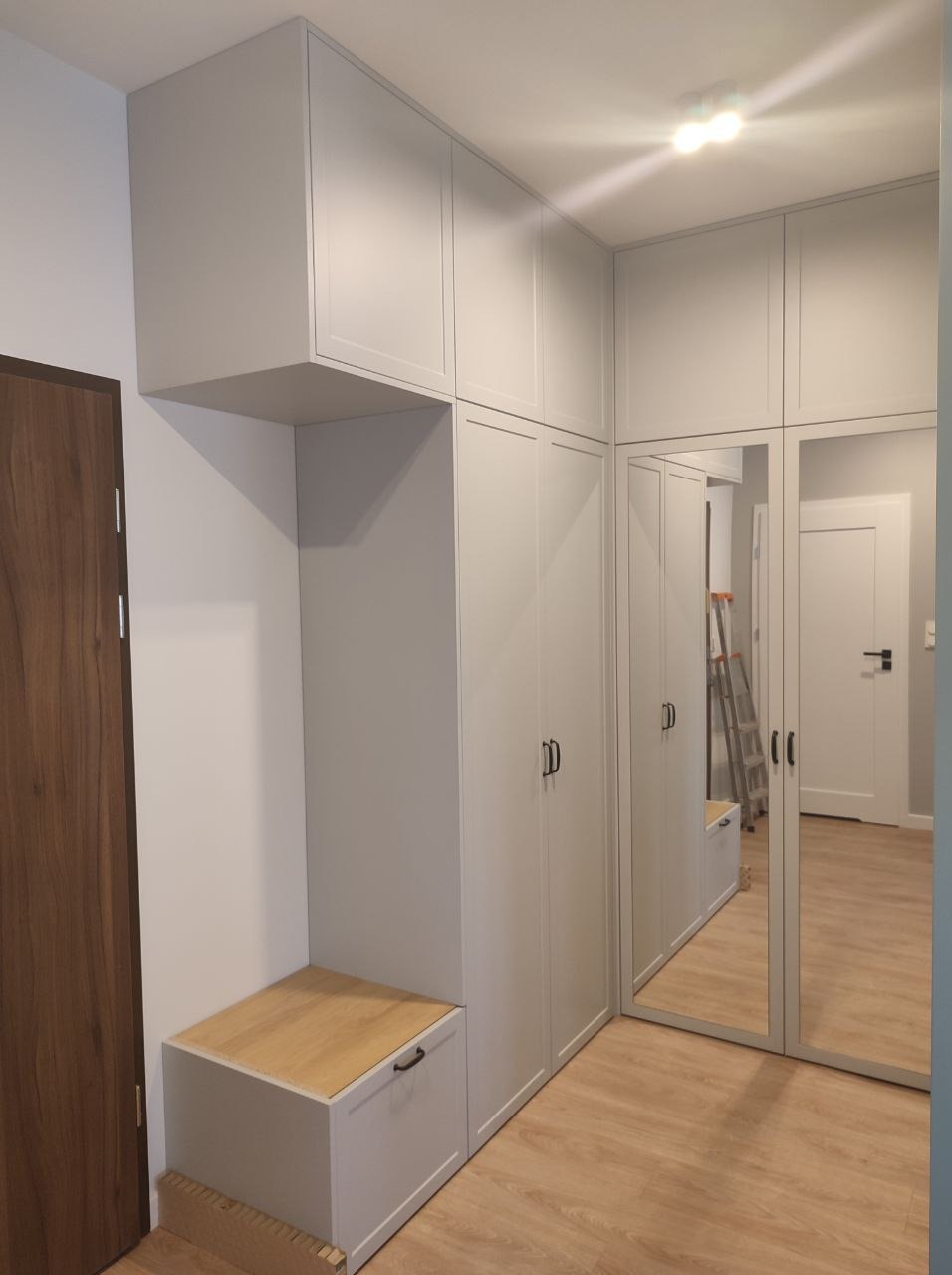 Jasnoszara szafa wnękowa z siedziskiem i lustrami, dopasowana do przedpokoju. Minimalistyczny design, drewniana podłoga, widoczne drzwi wejściowe.