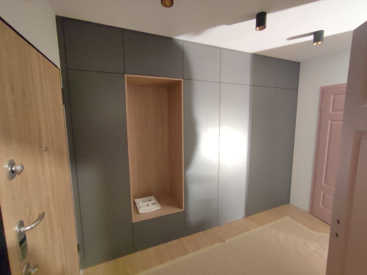 Szara szafa wnękowa z otwartą półką, widok z perspektywy wejścia. Minimalistyczny design, jasne drewno podłogi i drzwi. Nowoczesne oświetlenie sufitowe.