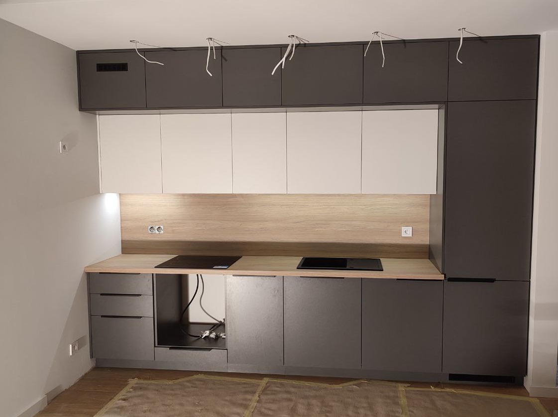 Nowoczesna kuchnia z szarymi i białymi szafkami, drewnianym blatem i płytą indukcyjną. Widoczne kable wystające z sufitu, sugerujące prace wykończeniowe.