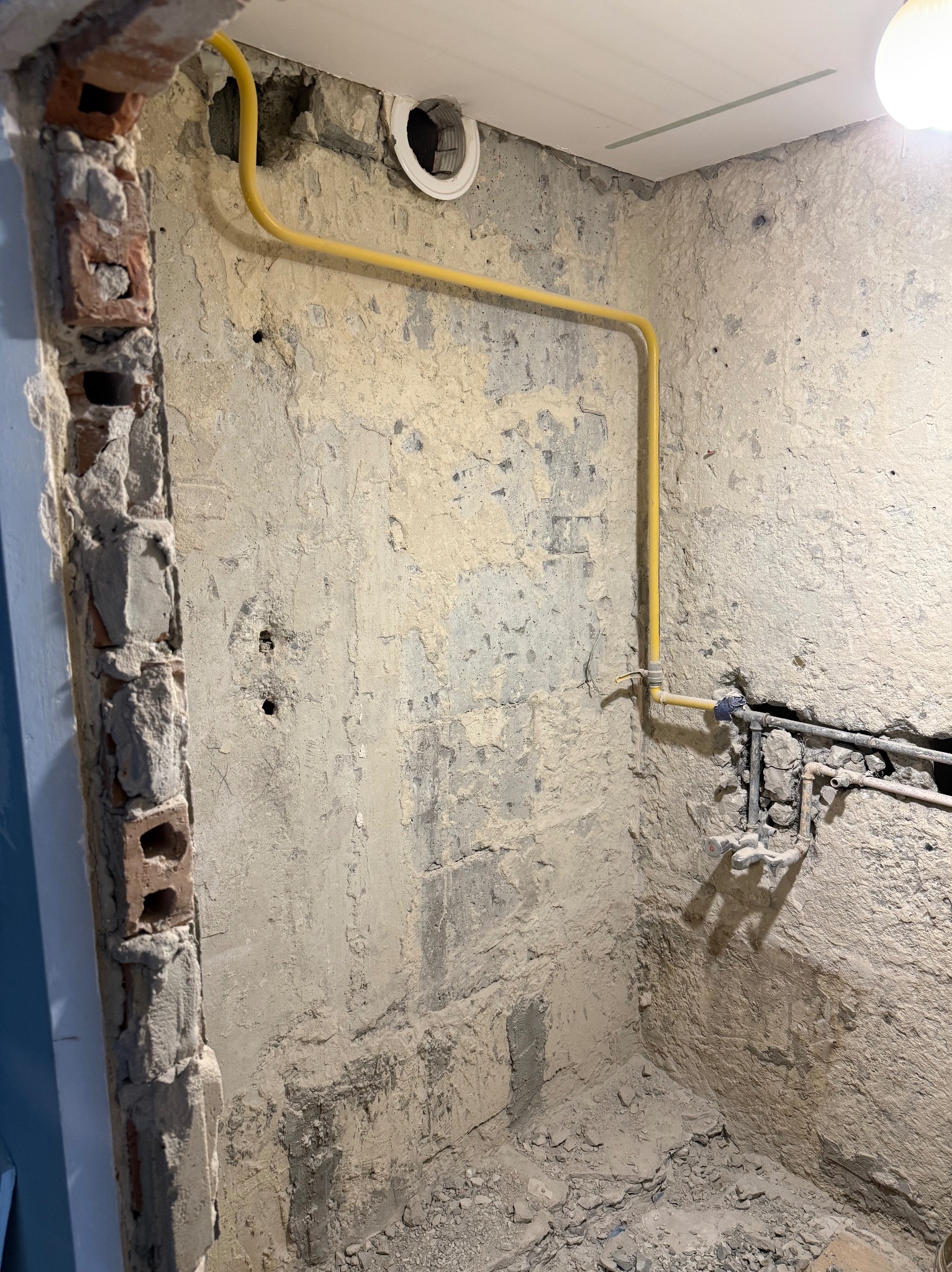 Surowa ściana w trakcie remontu łazienki z widoczną instalacją hydrauliczną i fragmentem żółtej rury gazowej. Widoczne ślady po skuciu tynku i cegły.