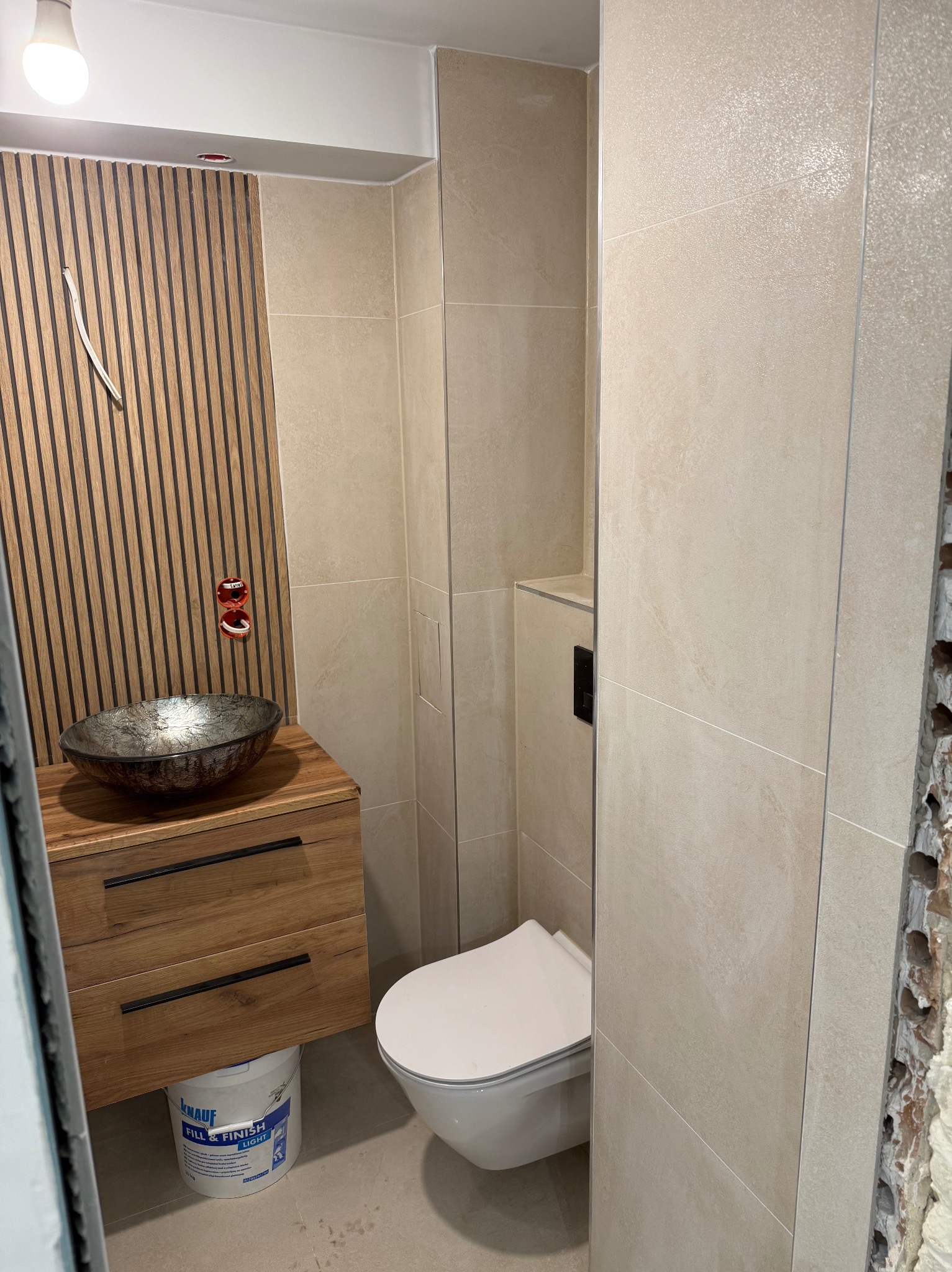 Nowoczesna łazienka z drewnianą szafką pod umywalkę i dekoracyjnymi lamelami na ścianie. Beżowe płytki i wisząca toaleta. Widoczny fragment ściany w trakcie wykończenia.