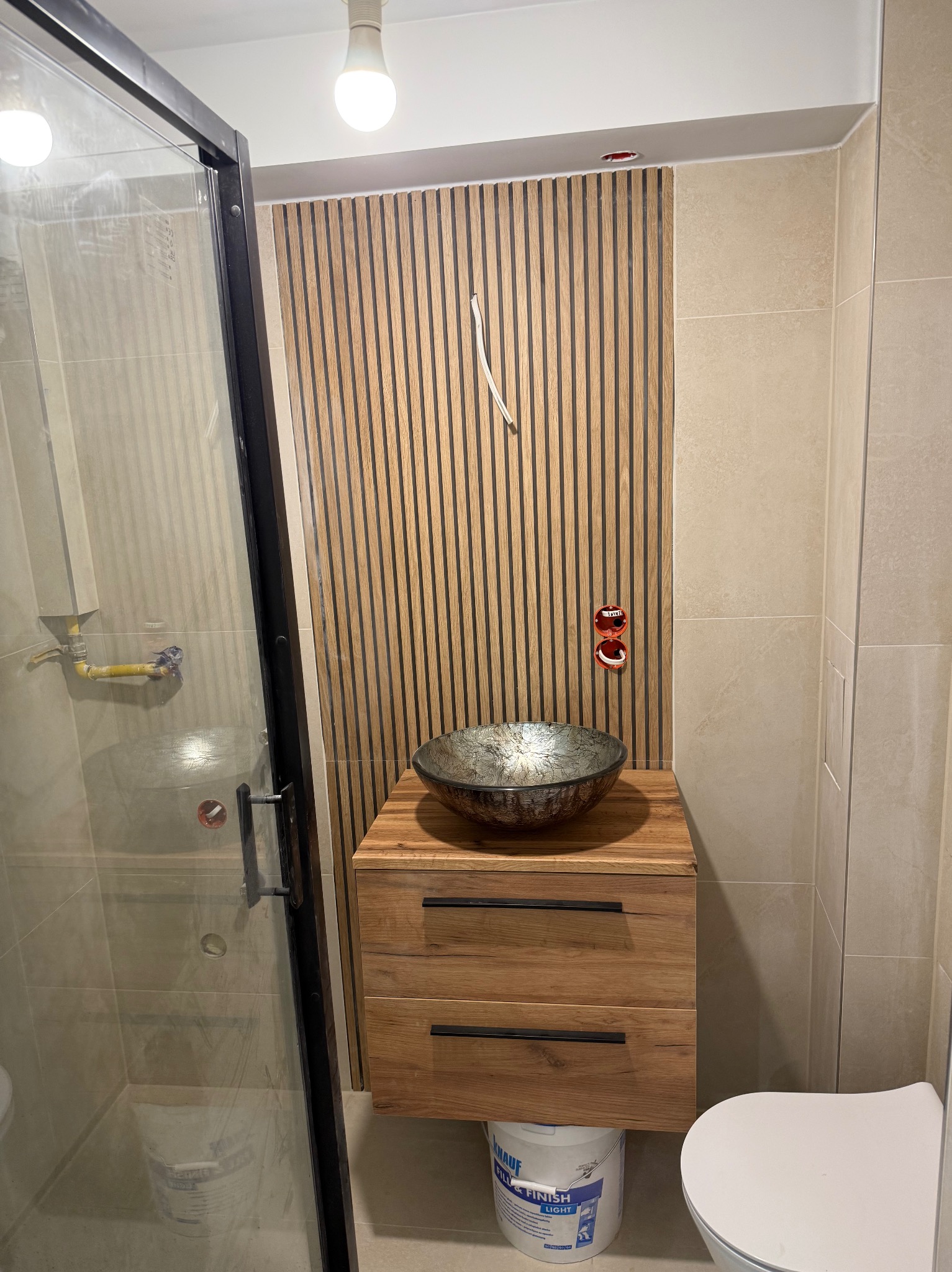 Wykończona łazienka z kabiną prysznicową, drewnianą szafką z umywalką i panelami ściennymi. Widoczne detale instalacji elektrycznej i sanitarnej.