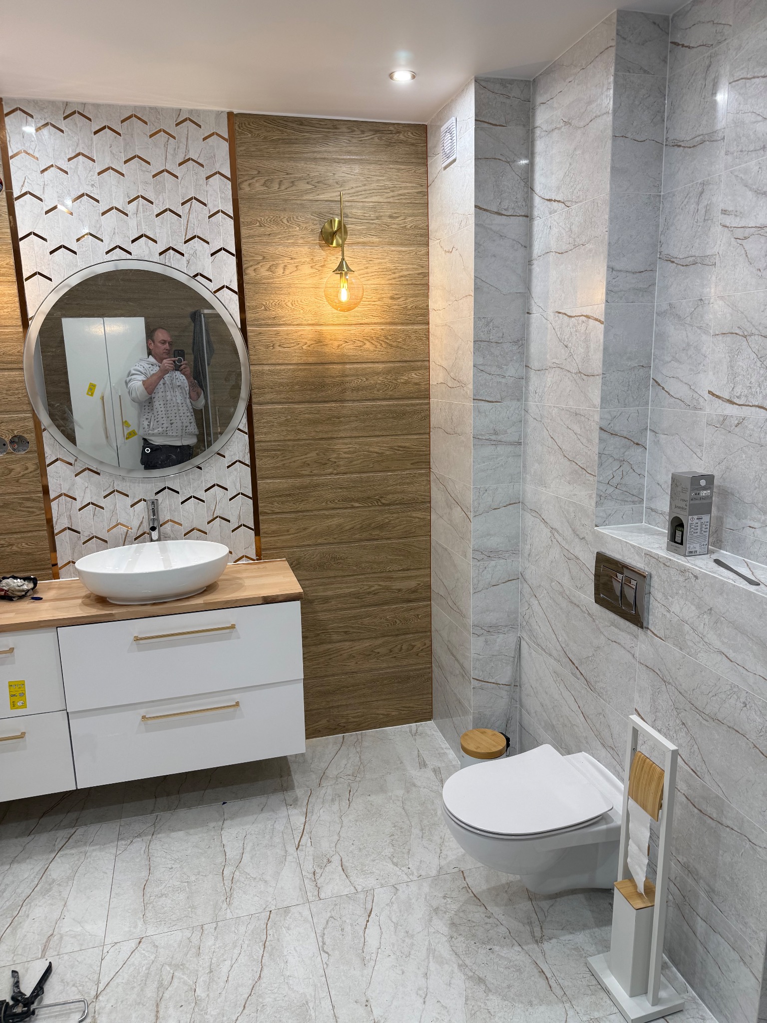 Nowoczesna łazienka po remoncie: biała umywalka na szafce, toaleta podwieszana, płytki imitujące marmur i drewno, okrągłe lustro z odbiciem osoby.