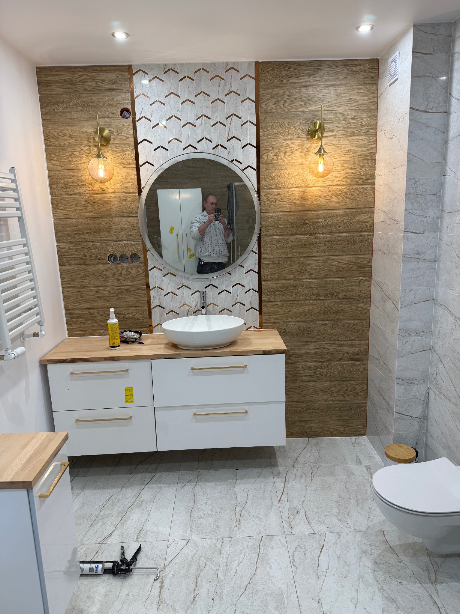 Nowoczesna łazienka po remoncie: drewnopodobne panele, mozaika w jodełkę, okrągłe lustro z odbiciem osoby, biała szafka z drewnianym blatem i złote lampy.