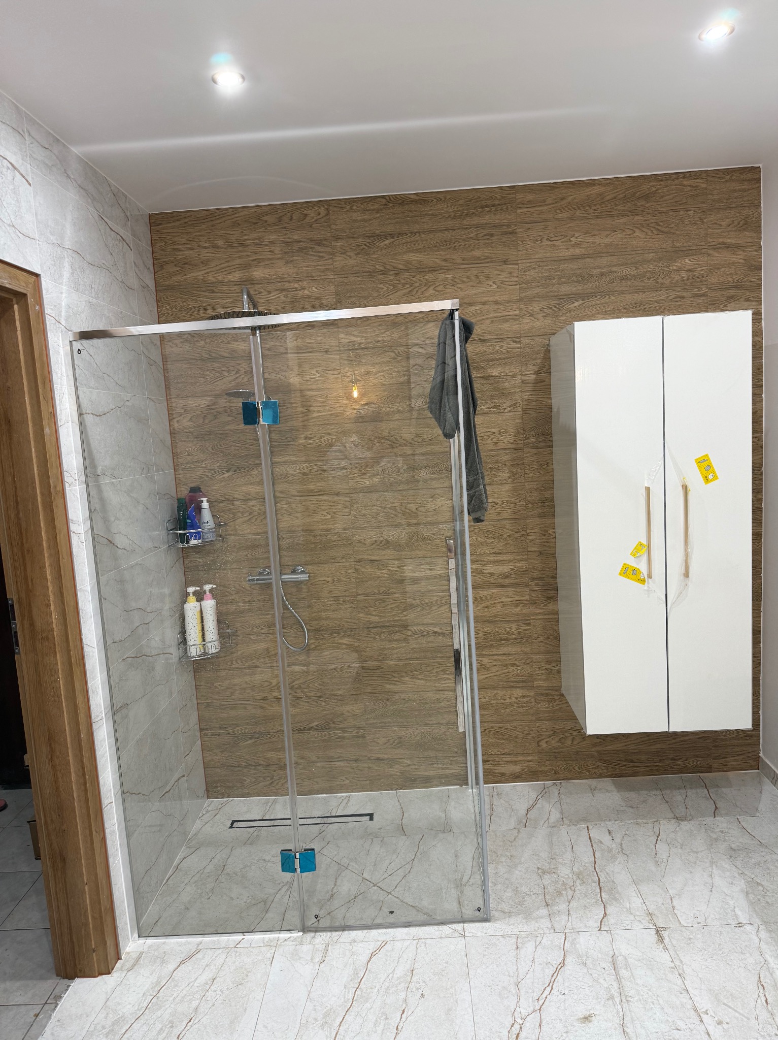 Nowoczesna kabina prysznicowa ze szklanymi drzwiami, drewnopodobne płytki na ścianie, biała szafka łazienkowa i marmurowe płytki podłogowe.