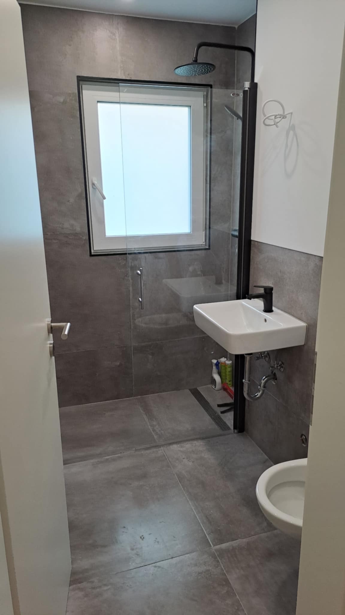 Nowoczesna łazienka z szarymi płytkami, kabiną prysznicową typu walk-in z czarną ramą, białą umywalką i częściowo widoczną toaletą. Minimalistyczny design.