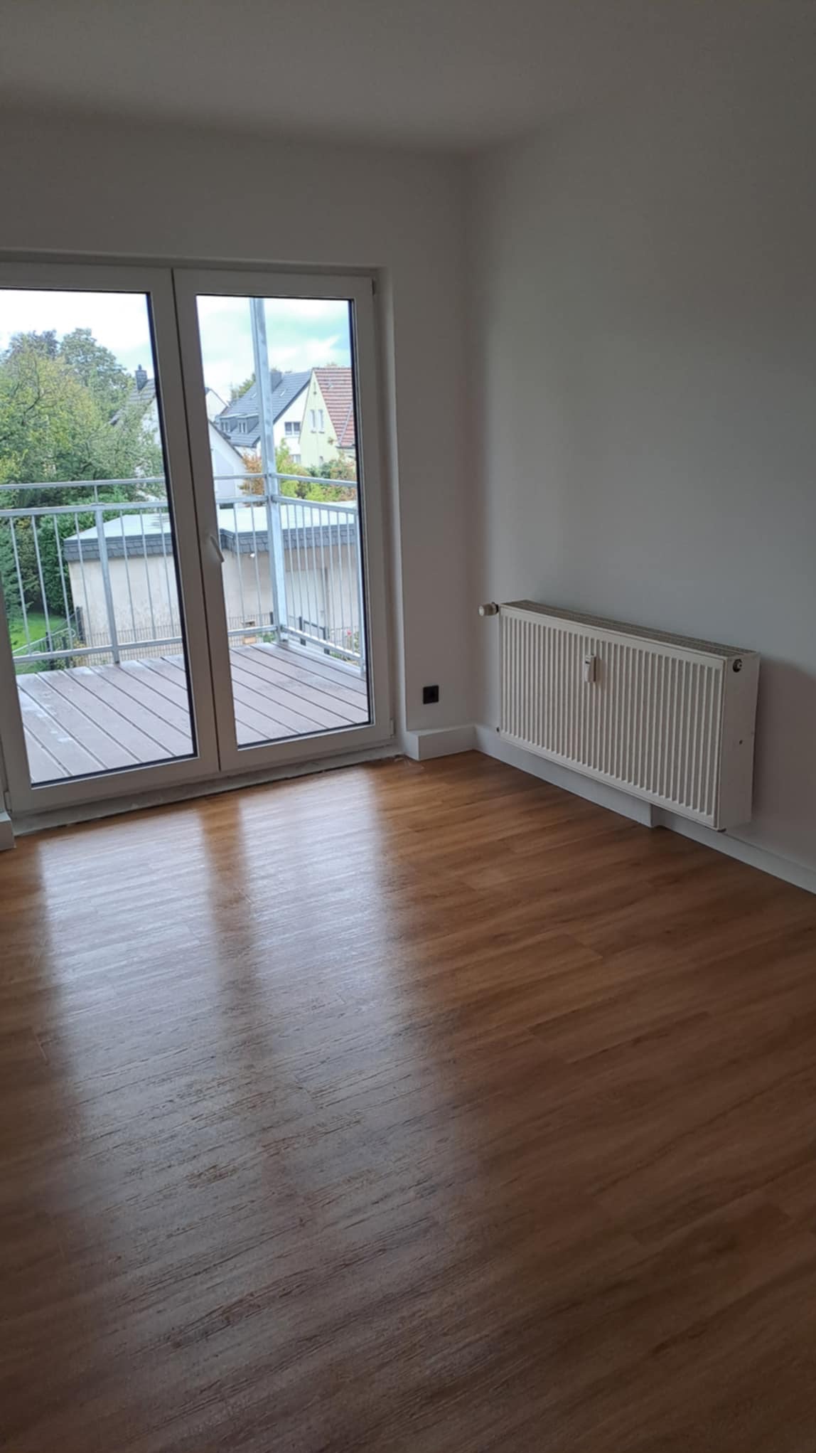 Jasne wnętrze pokoju z drewnianą podłogą, białymi ścianami, grzejnikiem i wyjściem na balkon z metalową barierką. Widok na zabudowania za balkonem.
