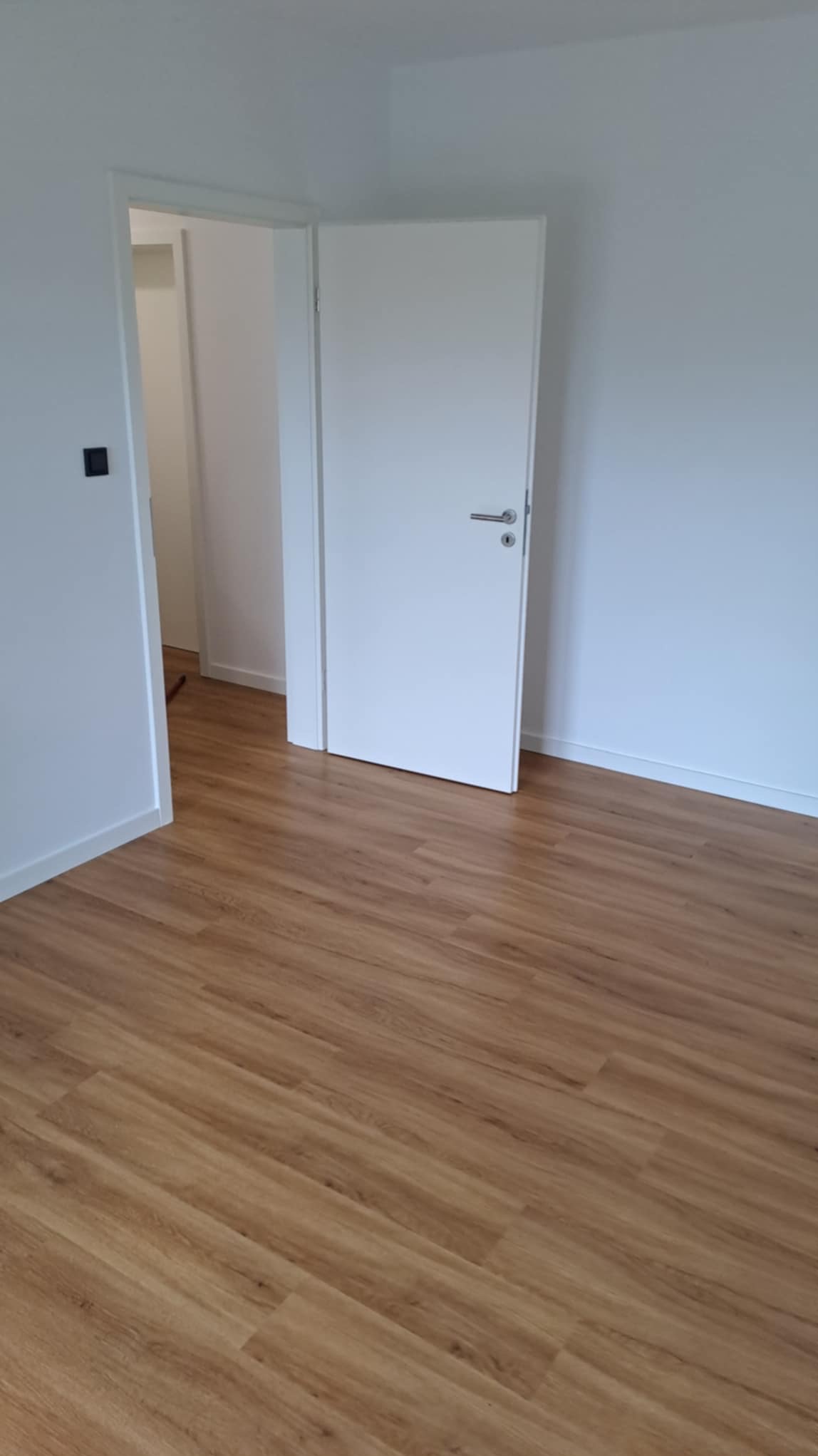 Jasne panele podłogowe imitujące drewno w nowym, minimalistycznym wnętrzu. Białe ściany i drzwi, widoczny fragment drugiego pomieszczenia.