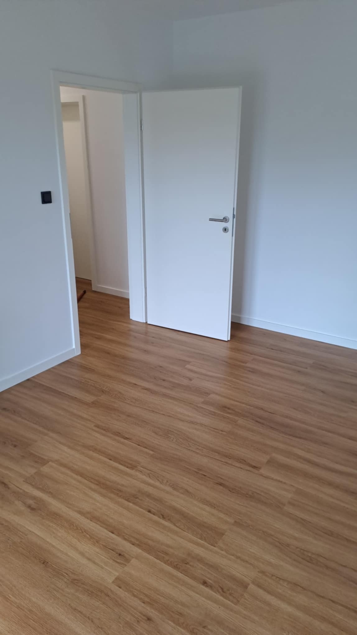 Jasne panele podłogowe w nowym, minimalistycznym wnętrzu z białymi ścianami i otwartymi drzwiami. Widoczny fragment włącznika na ścianie.