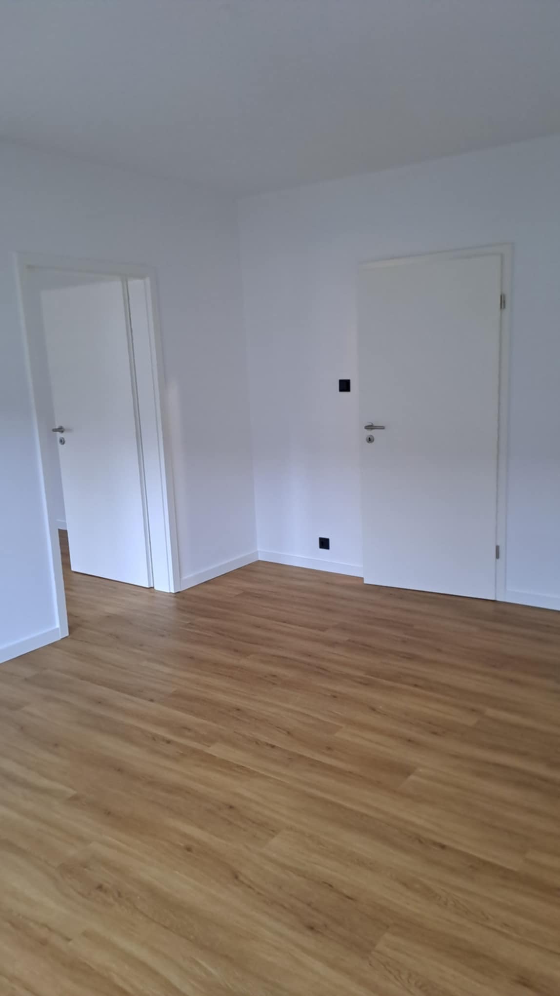 Jasne wnętrze po remoncie: panele podłogowe w odcieniach drewna, białe ściany z czarnymi włącznikami i dwoje białych drzwi z srebrnymi klamkami.