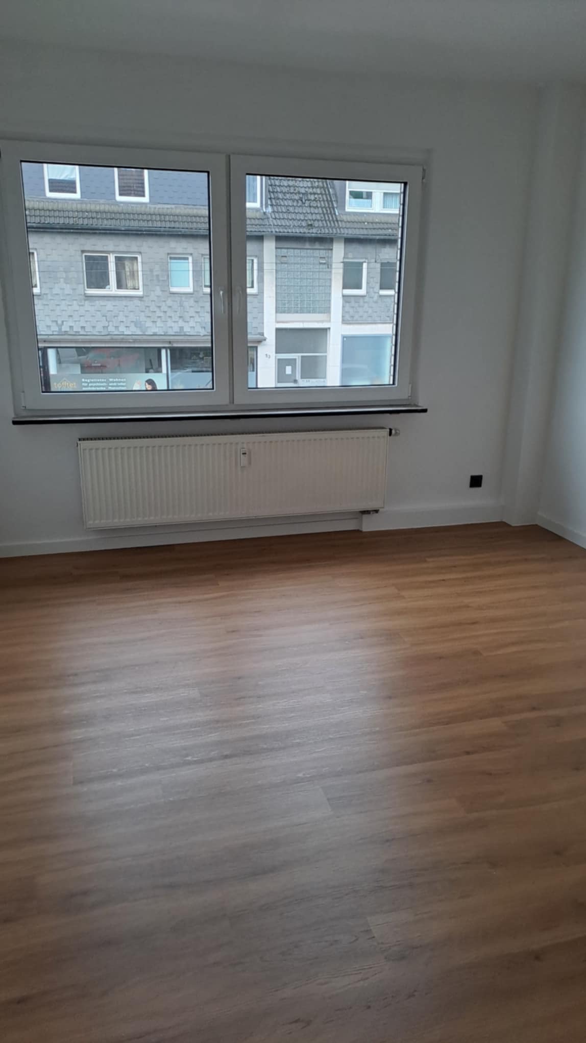 Jasne panele podłogowe w pokoju z białymi ścianami, grzejnikiem pod oknem i widokiem na budynki z zewnątrz. Minimalistyczne wnętrze po remoncie.