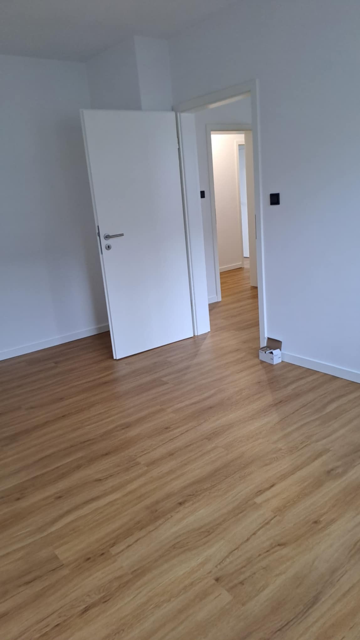 Jasne panele podłogowe w nowym, pustym pokoju z otwartymi białymi drzwiami i widokiem na kolejne pomieszczenia. Minimalistyczny design, świeżo po remoncie.