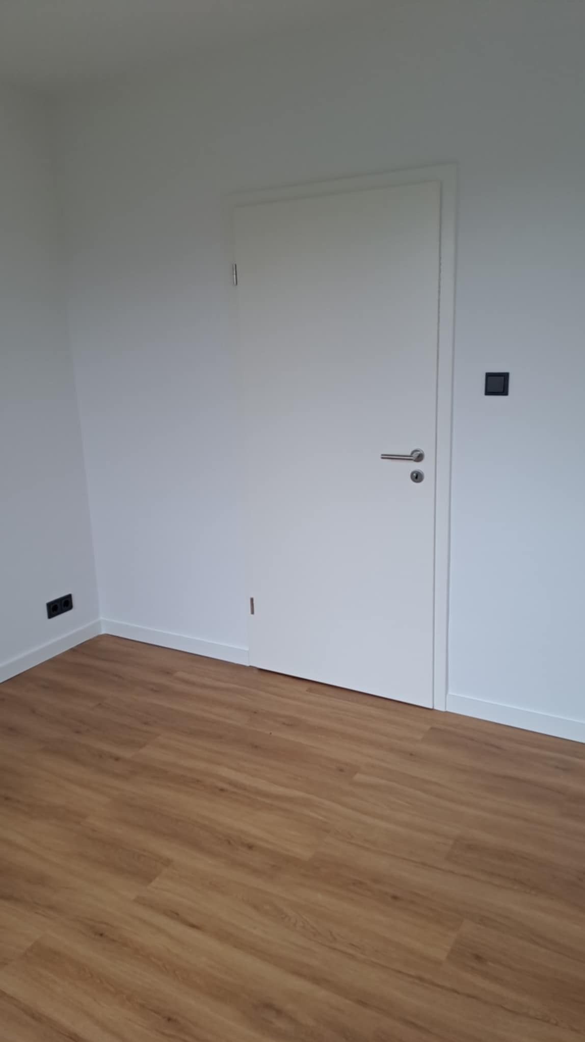 Jasne wnętrze po remoncie: panele podłogowe imitujące drewno, białe ściany z listwami przypodłogowymi i białe drzwi z srebrną klamką. Widoczne gniazdka elektryczne.