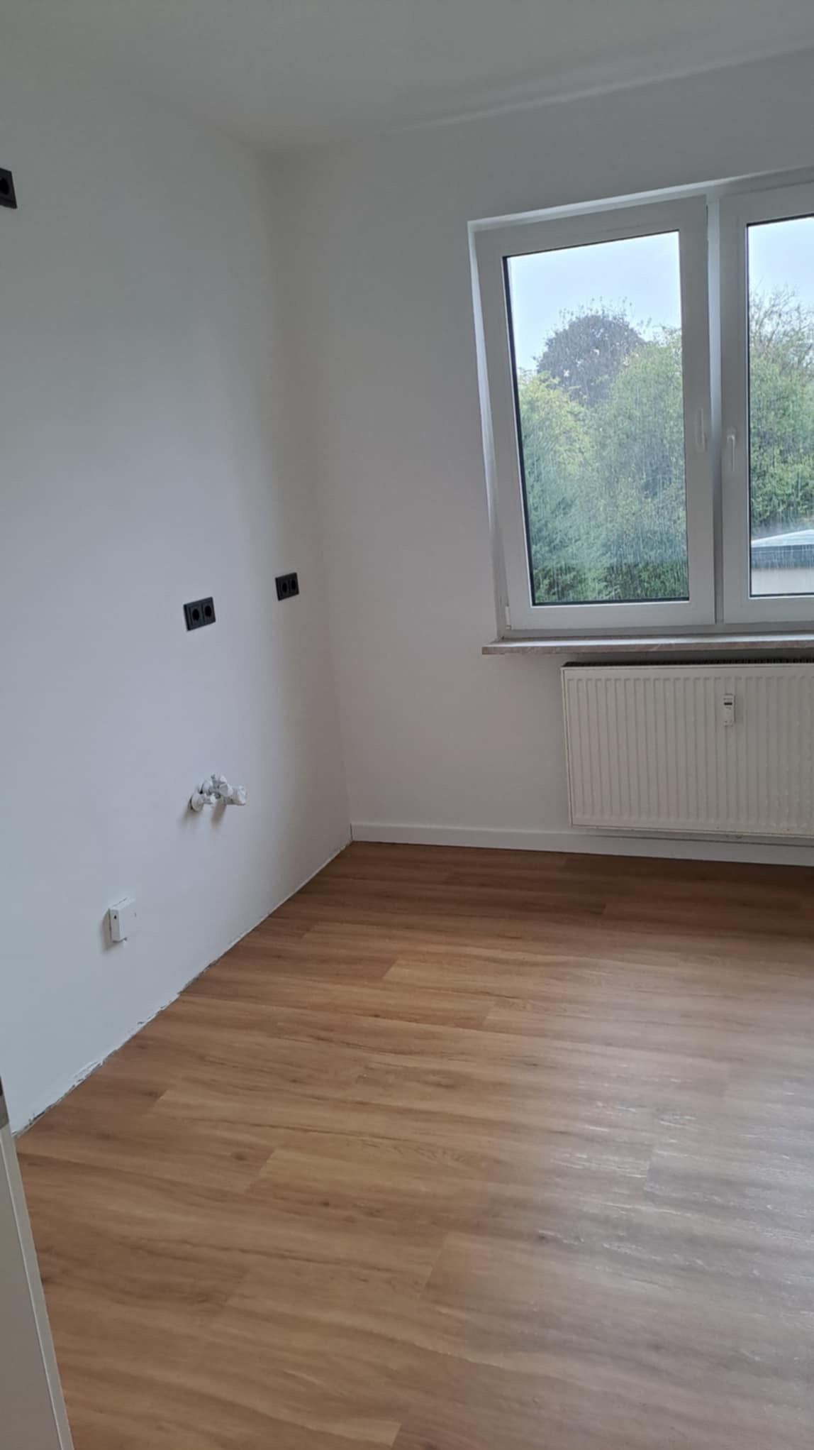 Jasne wnętrze po remoncie: panele podłogowe imitujące drewno, białe ściany z gniazdkami i grzejnik pod oknem. Widok na zieleń za oknem.