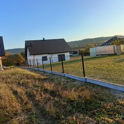 Bartłomiej Monica OGRO-TECH - Nowoczesne ogrodzenie panelowe z podmurówką betonową na nierównym terenie, w tle domy jednorodzinne i zalesione wzgórza w słoneczny dzień. Widok z dołu.