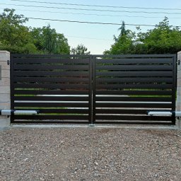 Bartłomiej Monica OGRO-TECH - Nowoczesna brama przesuwna z automatyką, poziome lamele w kolorze grafitowym, osadzona na słupkach z imitacji kamienia. Widoczne siłowniki i skrzynka na listy.