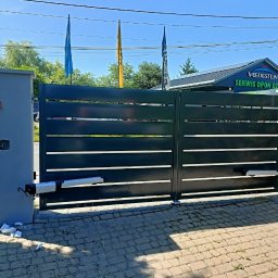 Bartłomiej Monica OGRO-TECH - Nowoczesna, pozioma brama metalowa w kolorze grafitowym z automatyką, zamontowana na betonowych słupkach. Widoczne okablowanie i fragmenty styropianu po montażu.