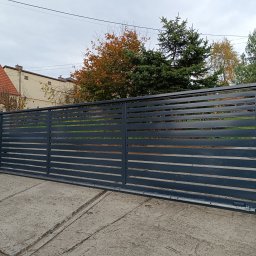 Bartłomiej Monica OGRO-TECH - Nowoczesna, przesuwna brama w kolorze antracytu z automatyką, zamontowana na posesji z betonowym ogrodzeniem i rzeźbą lwa. Widok z boku, jesienne drzewa w tle.